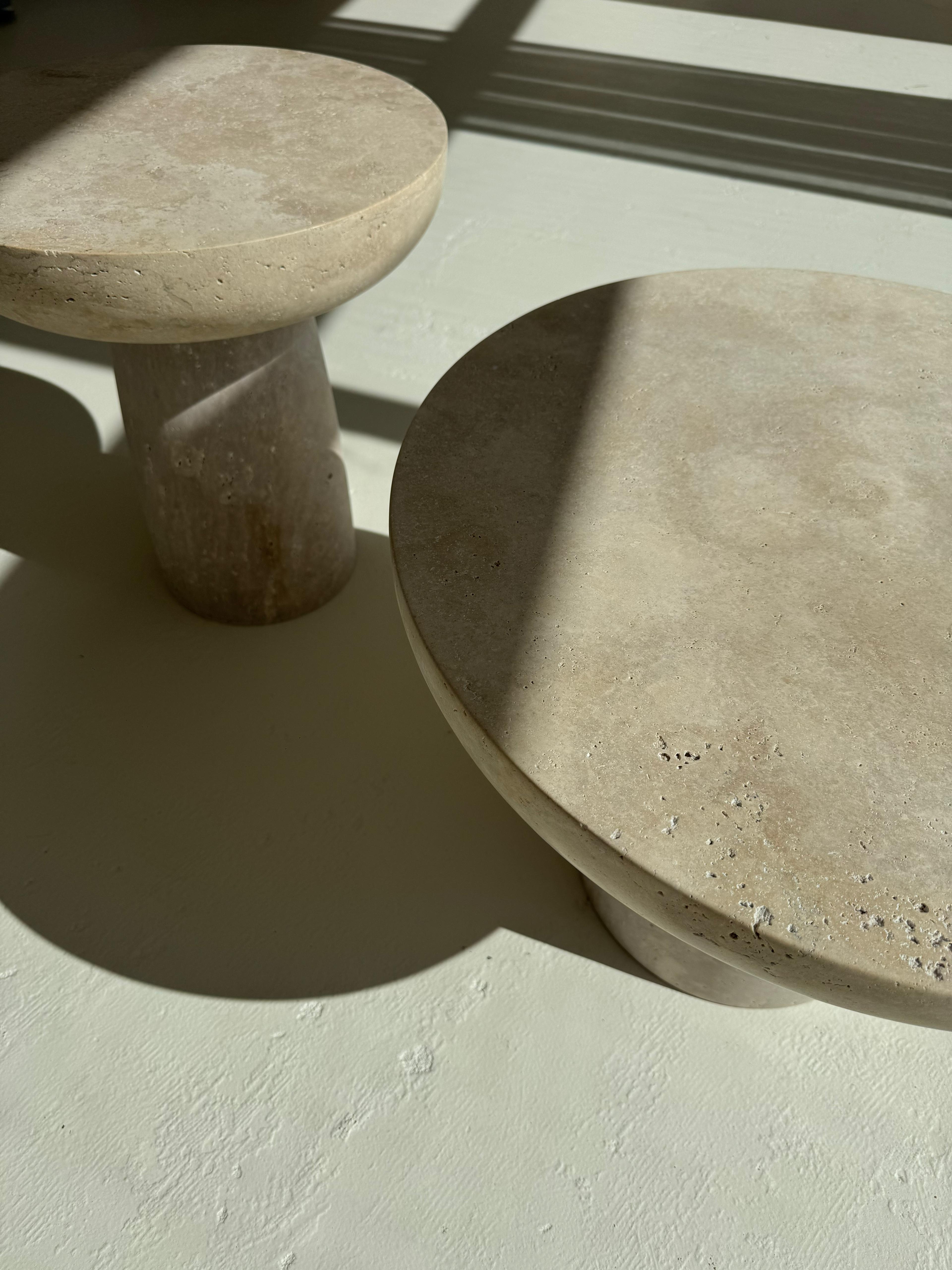 Set of two round travertine coffee tables with sculptural bases and polished stone tops in beige tones, styled in a modern neutral living room.