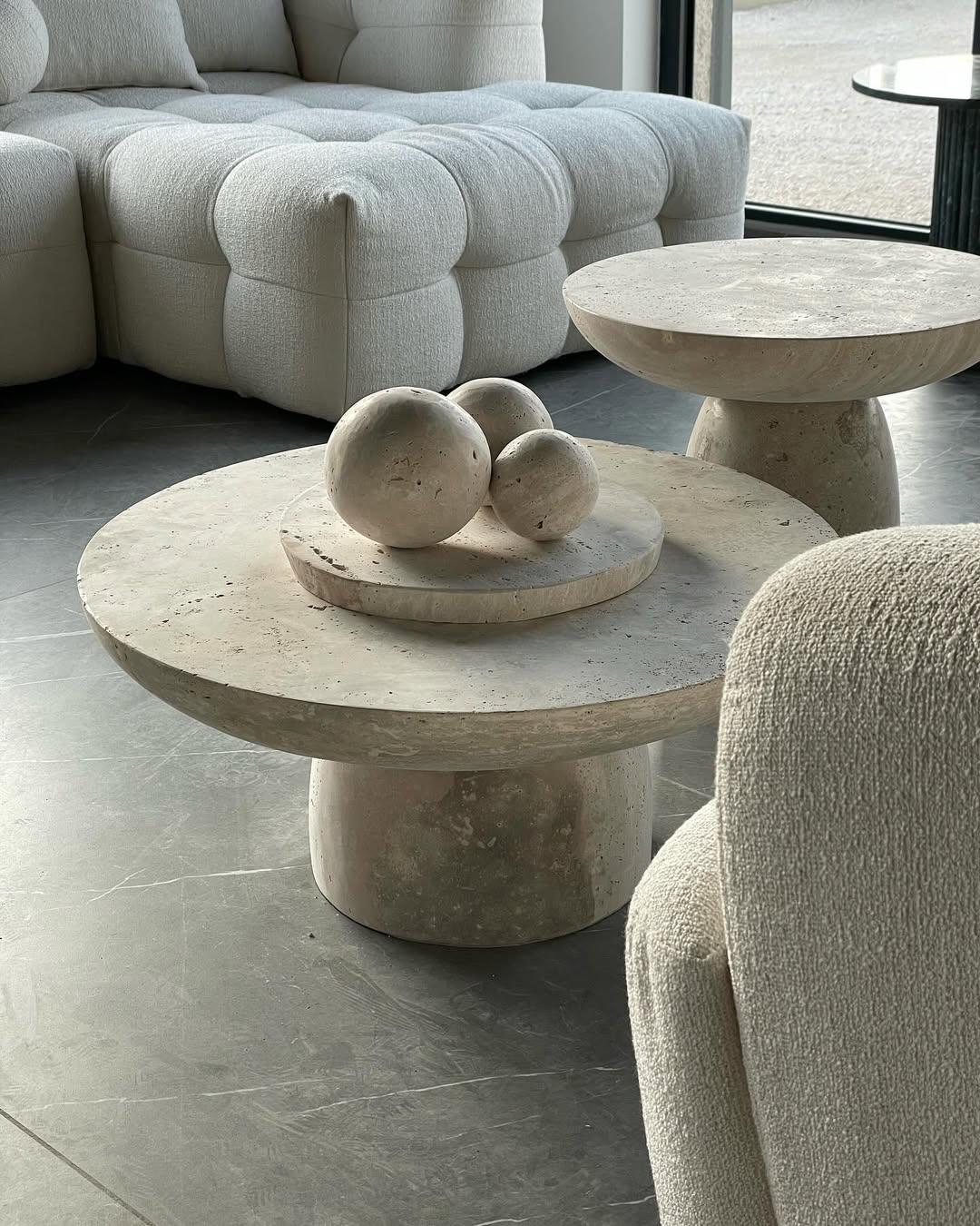 Set of two round travertine coffee tables with sculptural bases and polished stone tops in beige tones, styled in a modern neutral living room.