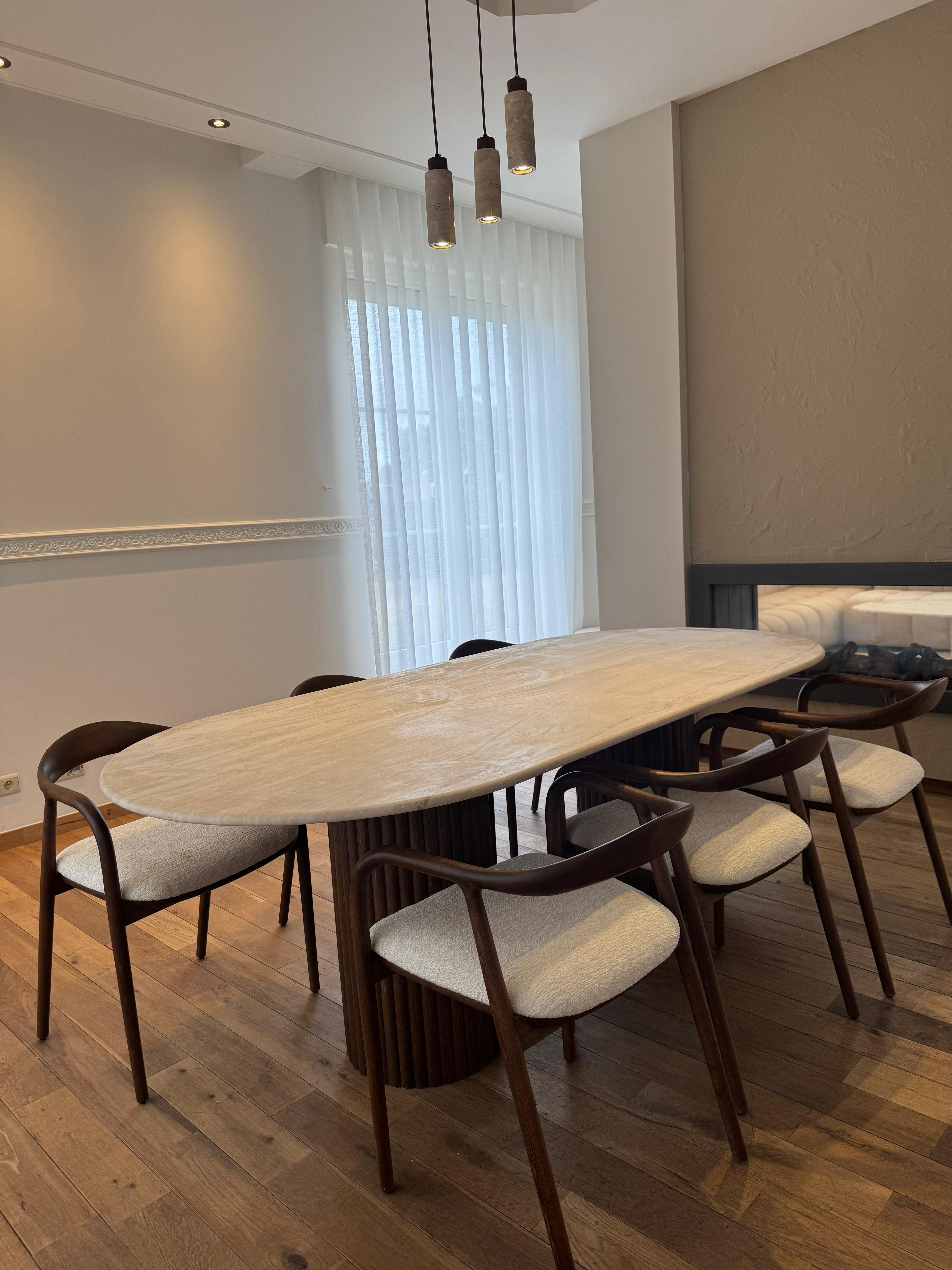Side view of the Vela | Travertine Oval Dining Table featuring ribbed walnut legs and a smooth travertine top in a softly lit interior.