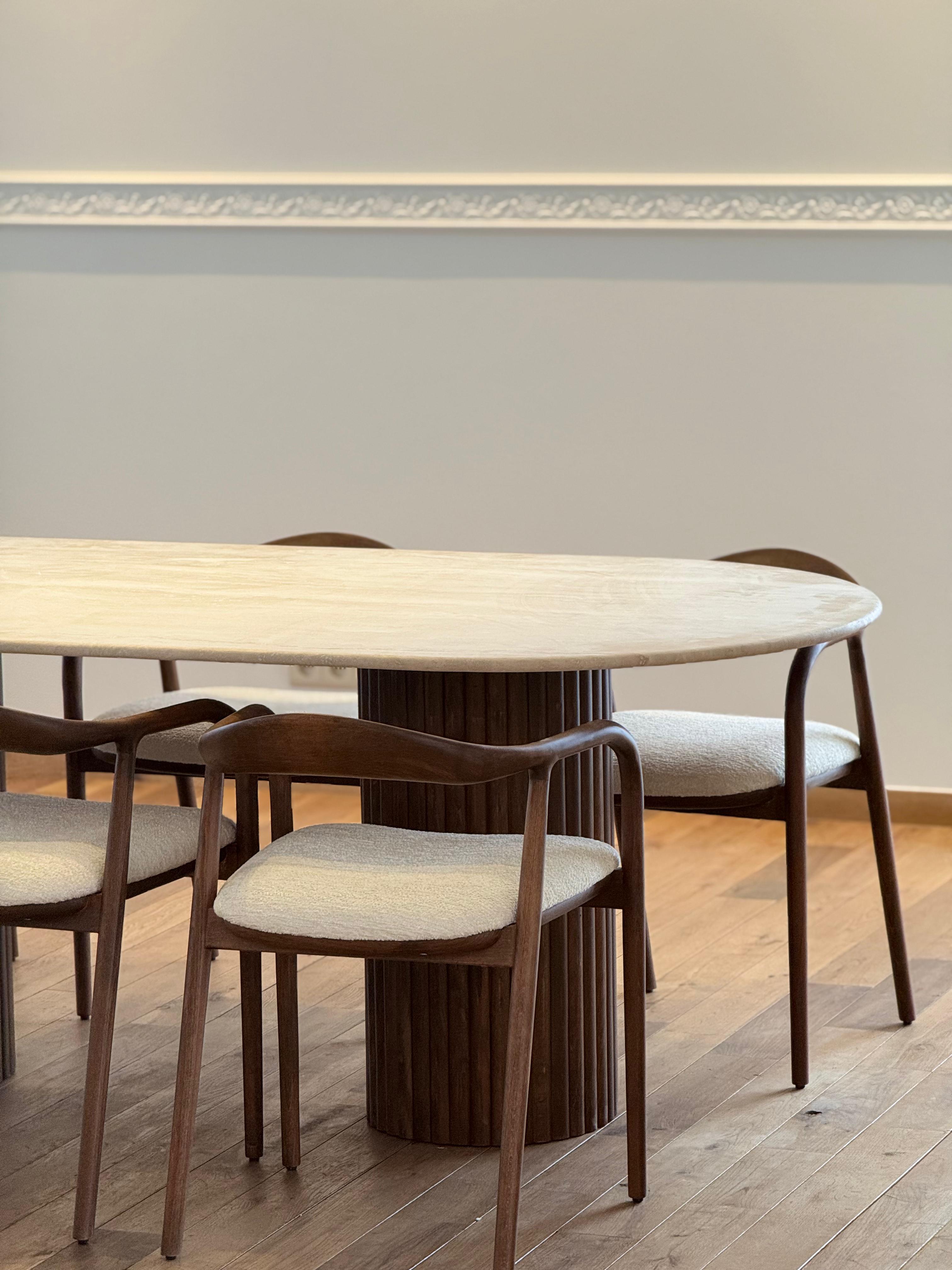 Side view of the Vela | Travertine Oval Dining Table featuring ribbed walnut legs and a smooth travertine top in a softly lit interior.