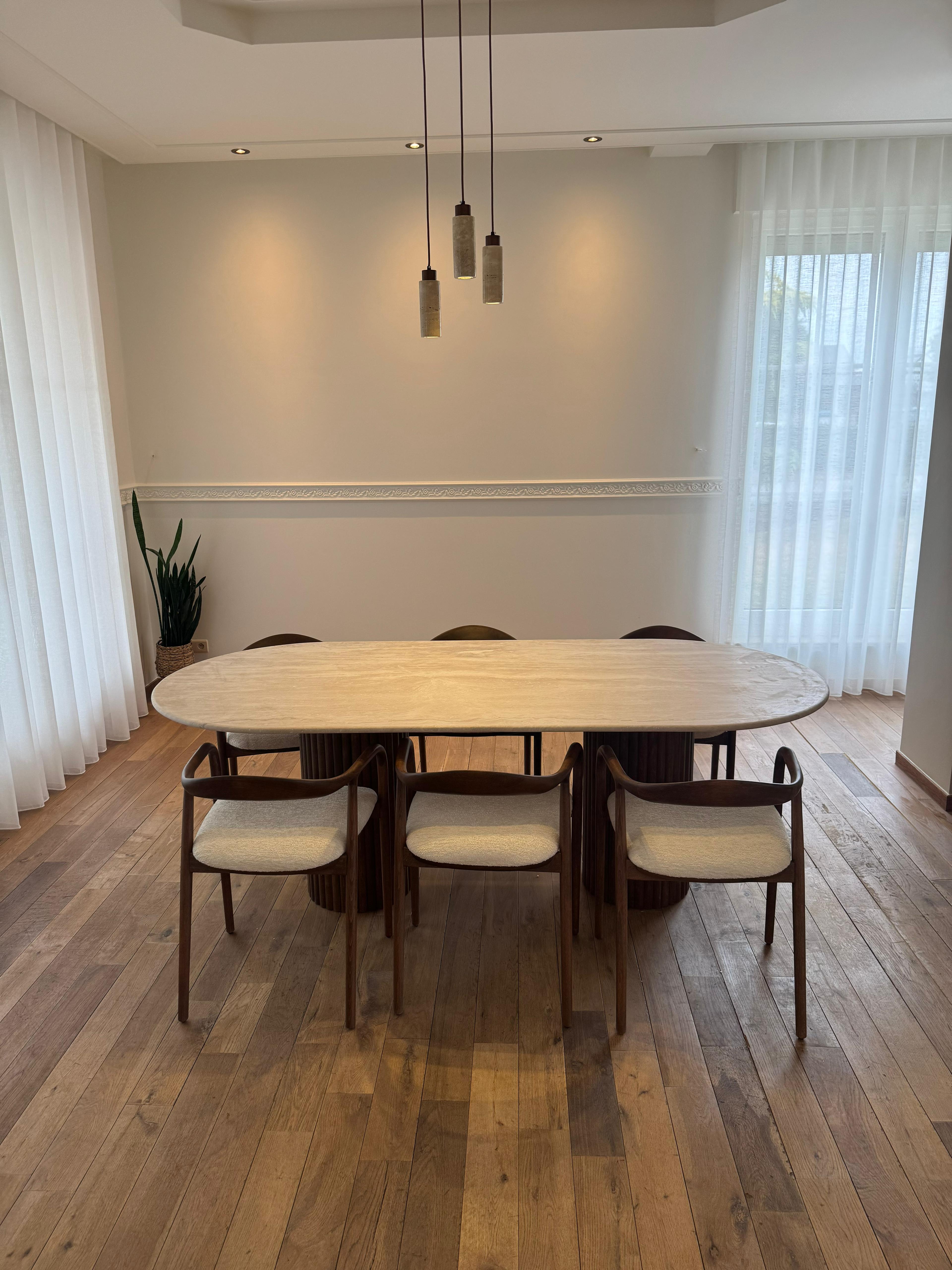 Side view of the Vela | Travertine Oval Dining Table featuring ribbed walnut legs and a smooth travertine top in a softly lit interior.