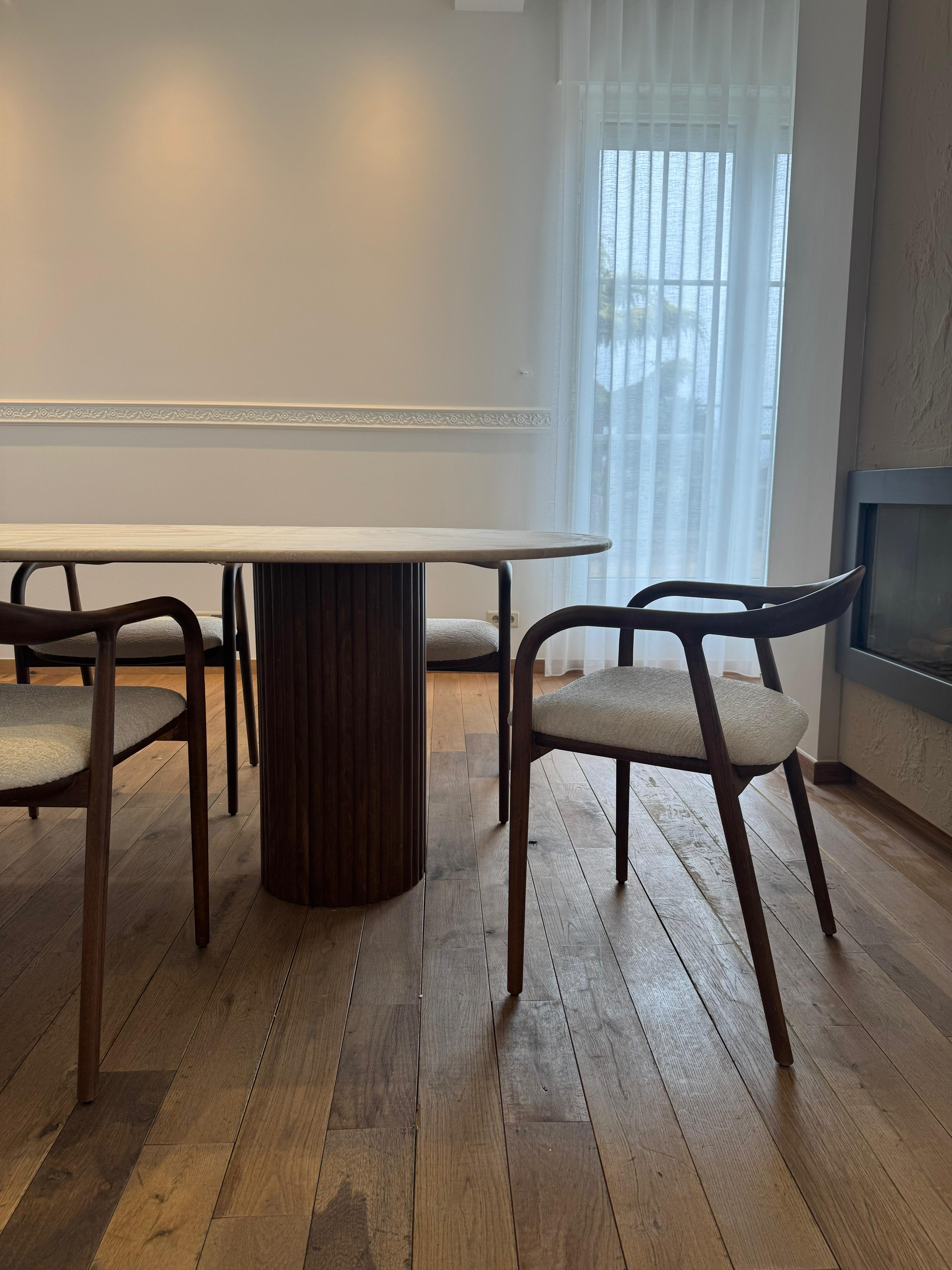 Side view of the Vela | Travertine Oval Dining Table featuring ribbed walnut legs and a smooth travertine top in a softly lit interior.
